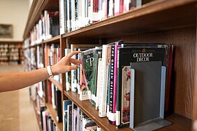 Zu sehen ist ein Bücherregal. Eine Hand zieht eines der Bücher aus dem Regal.